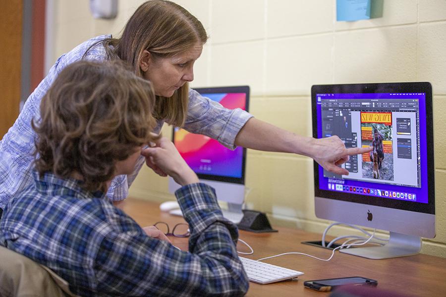 A faculty member points at a Mac computer with a student sitting in a chair in front of the computer.