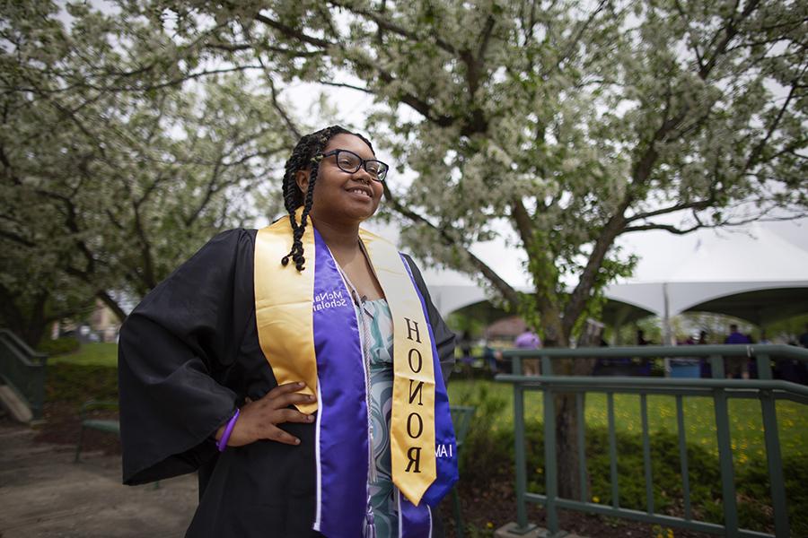 A student stands proudly in graduation attire with a yellow honors cord around her shoulders.